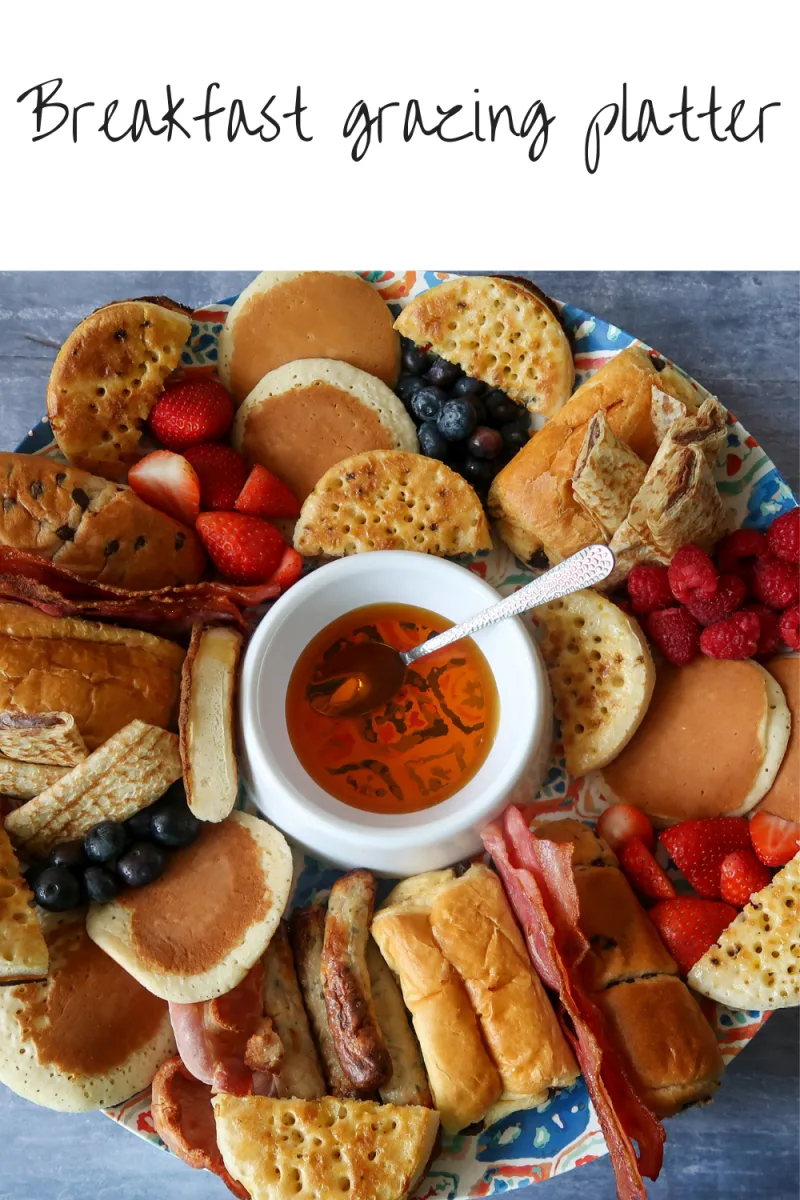 A breakfast platter containing crumpets, bacon, sausages, pain au chocolat, brioche rolls, fruit, and pancakes