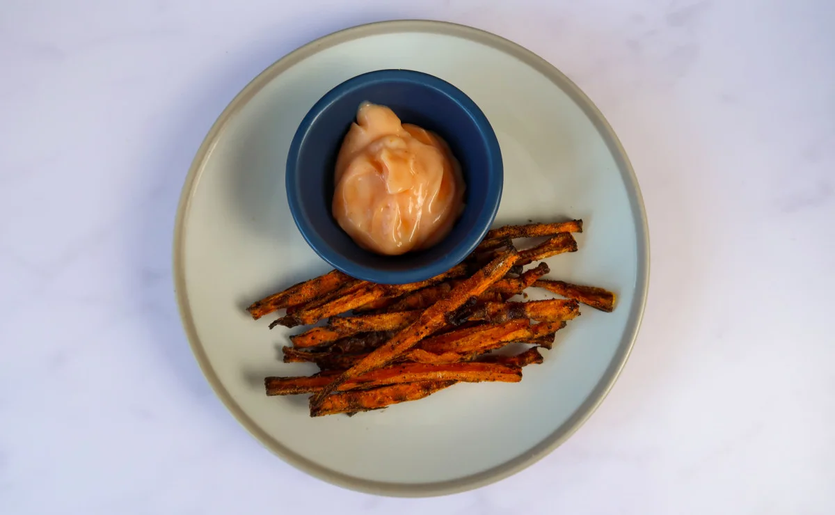 A white and mushroom coloured plate with cooked air fryer carrot fries with a blue circle bowl with sriracha mayonnaise.