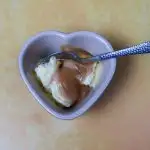 A heart shaped ramekin filled with vanilla ice cream and salted caramel sauce. There is a brushed aluminium teaspoon in the ice cream and it is set against a yellow backdrop.