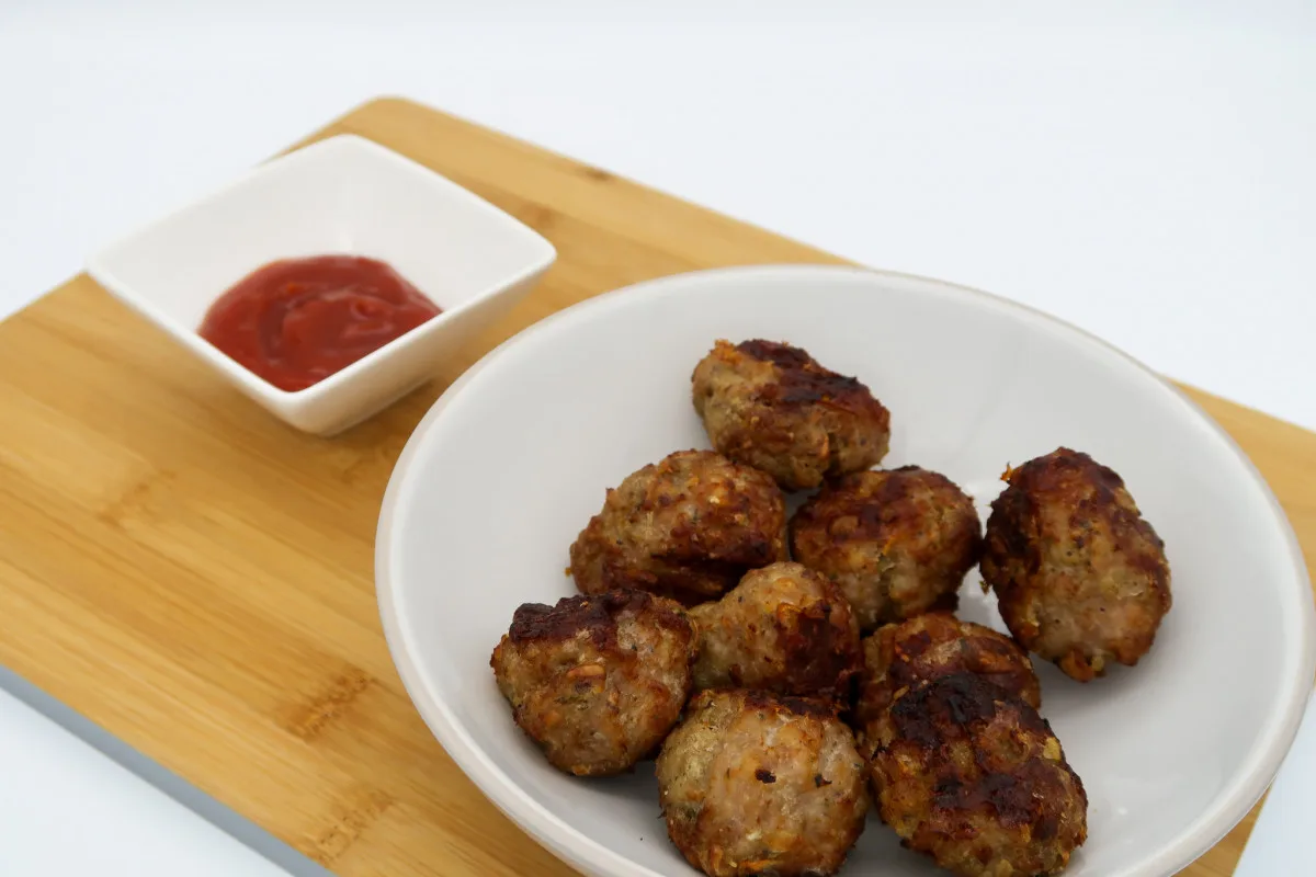 A white bowl filled with cooked air fryer sausage meatballs. The bowl is on a chopping board and in the background is a dish filled with tomato ketchup.