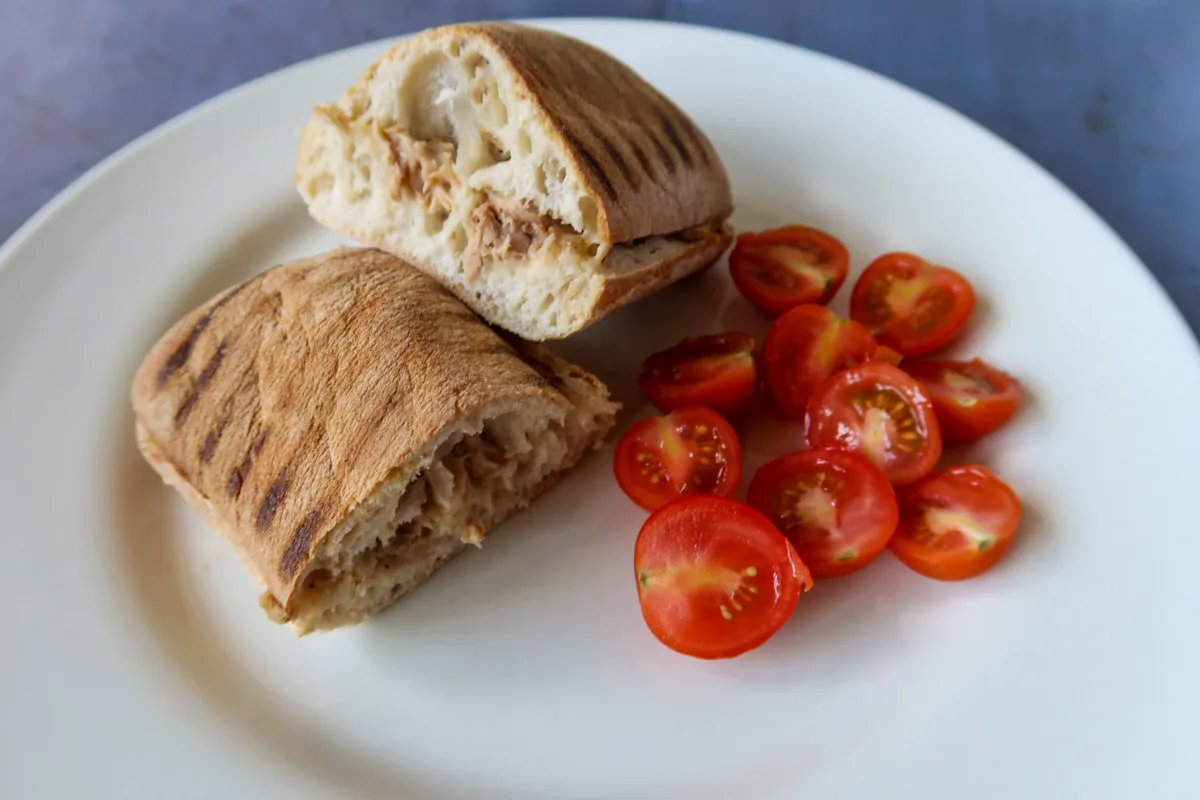 An air fryer tuna panini, cooked, cut in half, on a white plate, with halved cherry tomatoes on the side.