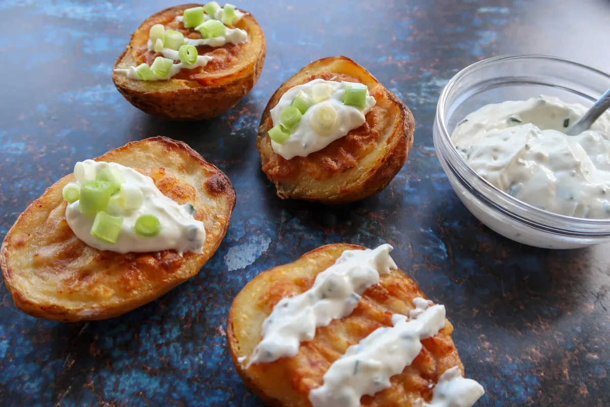 Cooked air fryer potato skins topped with sour cream and chive dip and spring onions. There is a bowl of sour cream and chive dip in the background also.