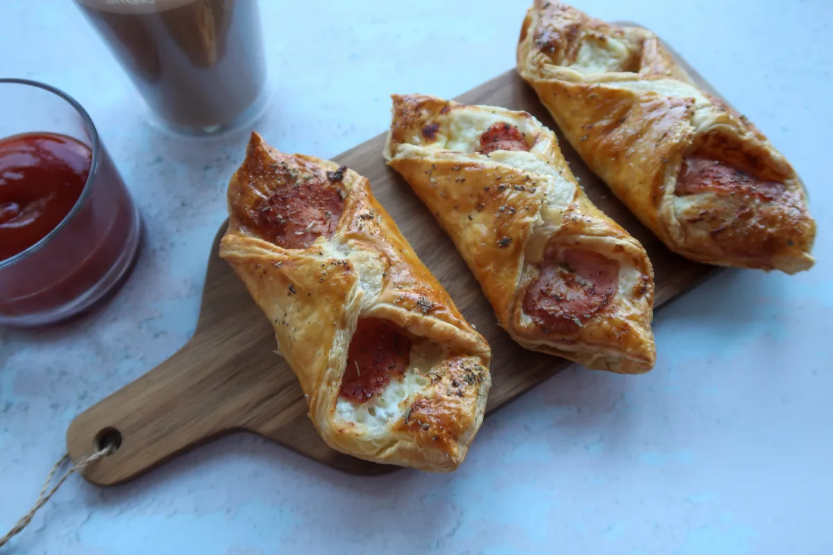 Three air fryer bacon and cheese turnovers on a chopping board. To the left of the board is a pot of tomato ketchup and a salted caramel latte.